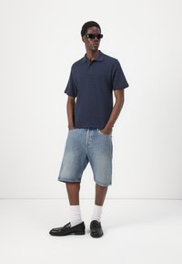 Mornarsko modra polo majica s teksturiranim vzorcem, svetlo modri jeans kratke hlače, črne čevlje in bele nogavice. Model stoji z rokami v žepih.