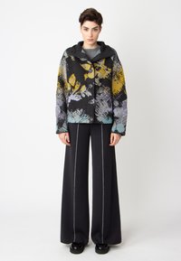Květinově vzorovaná bunda v černé, žluté a fialové barvě s kapucí a předními knoflíky, doplněná o široké tmavé kalhoty s bílým švem.