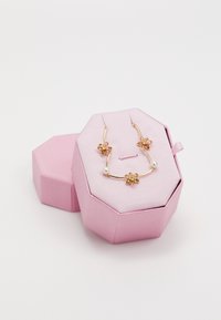 Collier floral en ton or avec des accents roses et des détails en perles présenté dans une boîte hexagonale rose clair à texture nervurée.