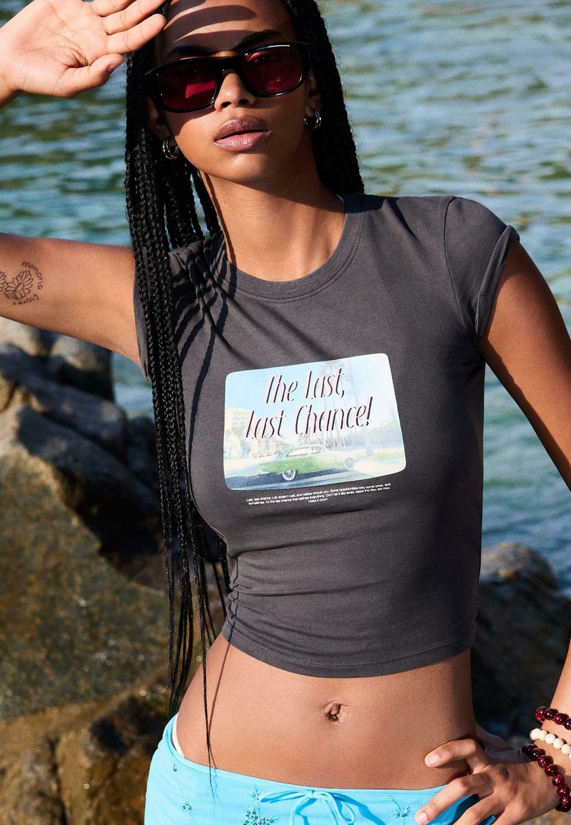 Sivý skrátený tričko s grafickým potlačou s nápisom "Posledná, posledná šanca!", na pozadí so scenériou a dizajnom vintage auta.