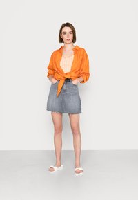 Camisa laranja de botão amarrada na cintura, top cropped xadrez, saia de ganga cinza com bainha desfiada e sandálias brancas com uma tira.