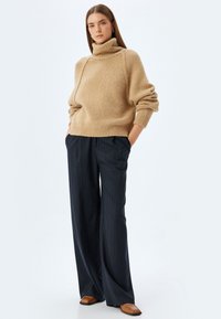 Maglione beige a collo alto lavorato a maglia con polsini a costine, abbinato a pantaloni a gamba larga navy con righe sottili e scarpe basse marroni.