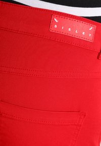 Červené bavlněné kalhoty s hladkou texturou, s výraznou značkovou etiketou "SISLEY" v bílé na červeném pozadí a standardní zadní kapsou.