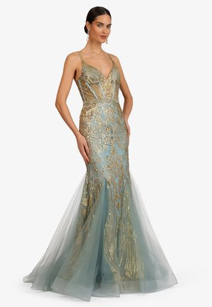 Frau trägt ein ärmelloses, blaues Meerjungfrauenkleid mit goldbesticktem, transparentem Tüllrock und dünnen Trägern, steht mit den Händen in der Hüfte.