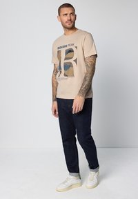 Beige bomulls-t-shirt med ett grafiskt tryck i blått och brunt, kombinerad med mörka jeans och vita sneakers med gröna detaljer.
