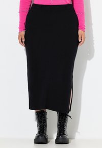 Vrouw in een zwarte midi-rok met een zij-split, zwarte veterschoenen voor de strijd, en een langwerpige roze top met franje, staande tegen een witte muur.
