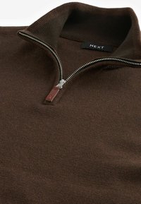 Brauner Reißverschluss-Pullover mit geripptem Kragen und Reißverschluss, der eine subtile Textur und ein Lederakzent am Reißverschluss zieht. Markenetikett sichtbar.