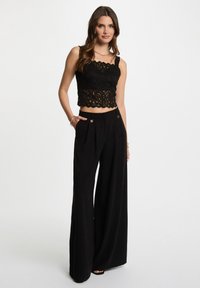 Top in pizzo nero corto con orlo a scallop abbinato a pantaloni neri a gamba larga a vita alta, caratterizzati da pence e dettagli metallici.