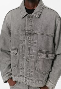 Osoba nosi izblijedjenu sivu traper jaknu s velikim prednjim džepovima i dugmadi, ruke dijelom u odgovarajućim sivim trapericama.