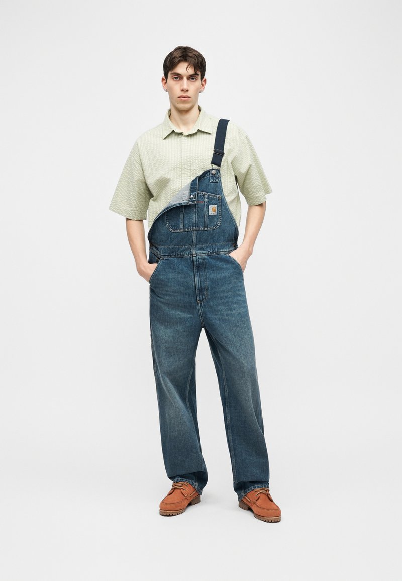 Junger Mann steht mit Händen in den Taschen, trägt ein hellgrünes kurzärmeliges Hemd, blaue Latzjeans mit einem Träger und braune Schuhe.