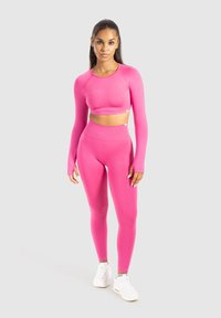 Vrouw staat rechtop in een felroze langemouwen crop top en hoge taille leggings, met witte sportschoenen, tegen een lichte achtergrond.
