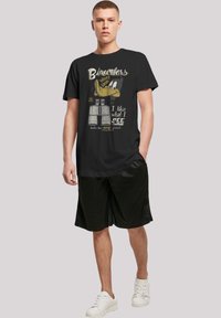 Svart bomull t-skjorte med grafikk av en tegneseriesangfugl og tekst. I kombinasjon med svarte shorts og hvite joggesko, står modellen foran en nøytral bakgrunn.