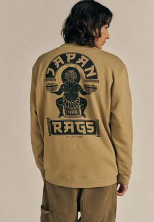 Persona che indossa una felpa beige con una grafica nera di un lottatore di sumo che tiene delle ciotole e le parole "Japan Rags" sulla schiena.