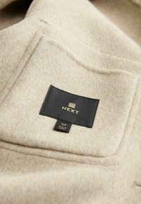 Gros plan sur la poche d’un manteau en laine beige avec une étiquette de marque NEXT noire et une étiquette de taille M cousue en dessous.