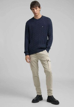 Suéter de punto de cable en azul marino, pantalones cargo beige con bolsillos laterales y botas negras. El suéter tiene cuello redondo y puños acanalados.