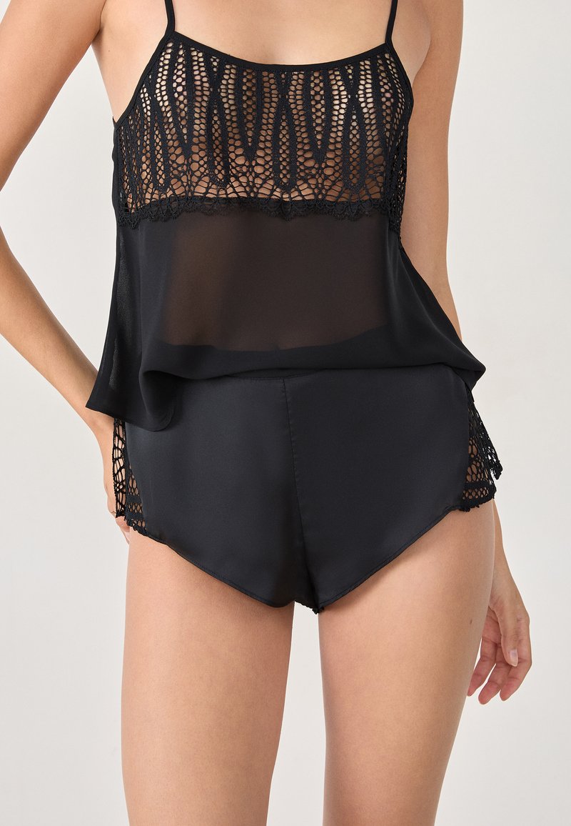 Modèle portant un haut noir transparent avec des détails en dentelle et un short assorti, se tenant devant un fond uni.