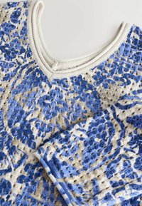 Strickpullover mit einer Mischung aus blauen Blumenmustern auf einem beigen Hintergrund, mit einem cremefarbenen gerippten Halsausschnitt und einer strukturierten Oberfläche.