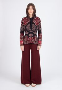 Blazer nero con intricati ricami floreali rossi e beige, abbinato a pantaloni larghi color bordeaux e scarpe con plateau beige.