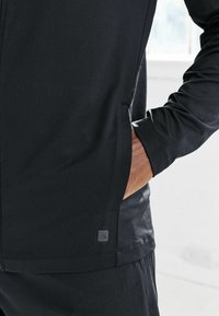 Musta urheilutakki, jossa on sileä pinta, vetoketjulliset sivut ja painettu logo helmassa. Hihansuut ovat istuvat.