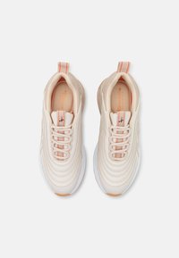 Light beige athletic shoes with textured, wavy overlays. Features white soles, orange accents, and flat laces. Loop tab on the heel.