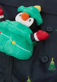 Ciemnogranatowa tkanina z zielonymi choinkami oraz pluszowa zabawka pingwina ubrana w zielony strój, z czerwonymi rękawiczkami i pomarańczowym dziobem.
