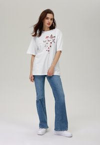 Hvit oversized T-skjorte med et sommerfugltrykk i forskjellige nyanser av rosa, kombinert med lyseblå, utsvingte denimjeans og hvite sneakers.