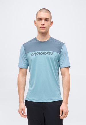 Mees, kes kannab hele- ja tumelsinist Dynafiti lühikeste varrukatega särki, seisab ühevärvilise valge tausta ees.