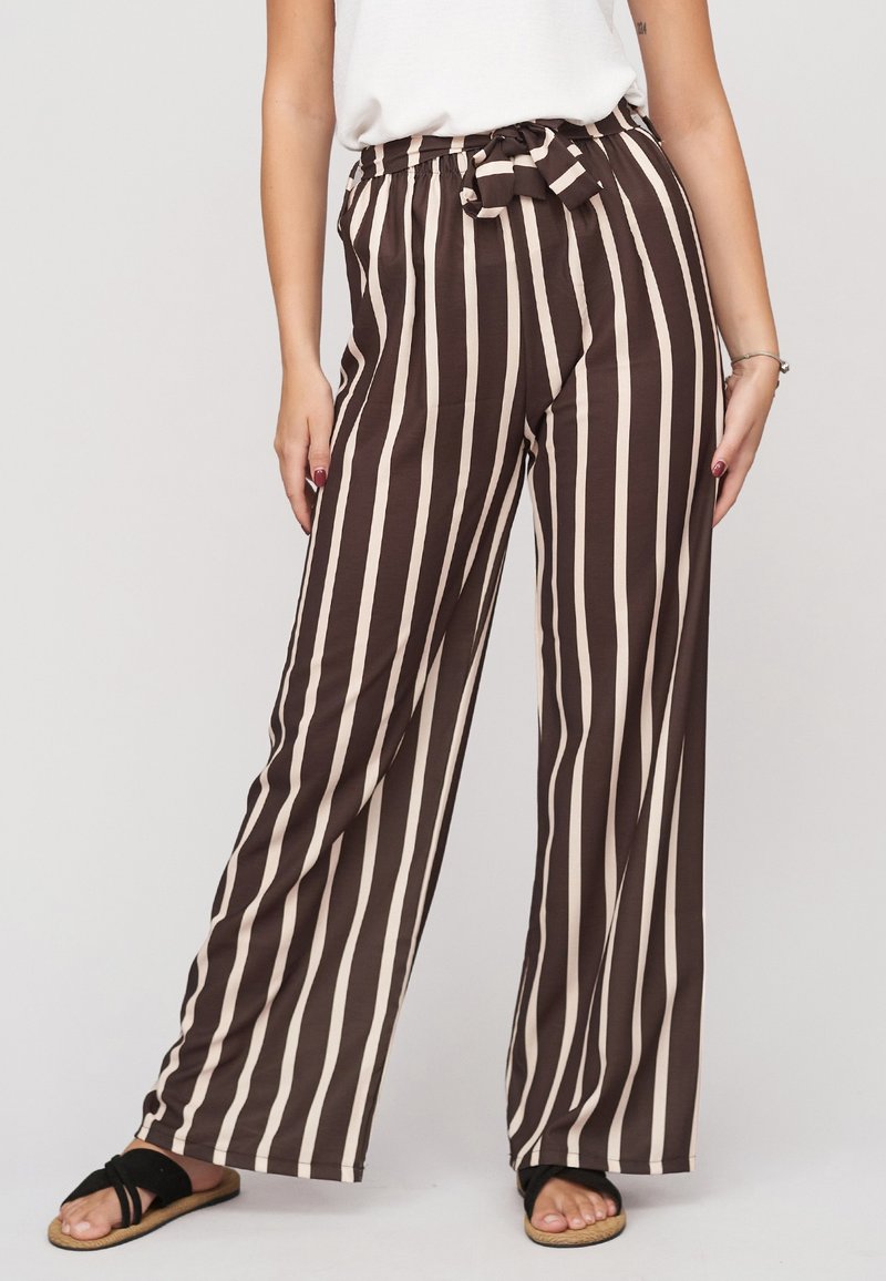 Femme portant un pantalon large à rayures verticales marron et blanc avec une taille nouée, haut blanc, sandales noires et bracelet argenté.