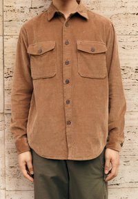 Brun kordfløjls-skjorte med to brystlommer, knaplukning foran og spids krave. Lange ærmer, ribbet tekstur og rundet kant.