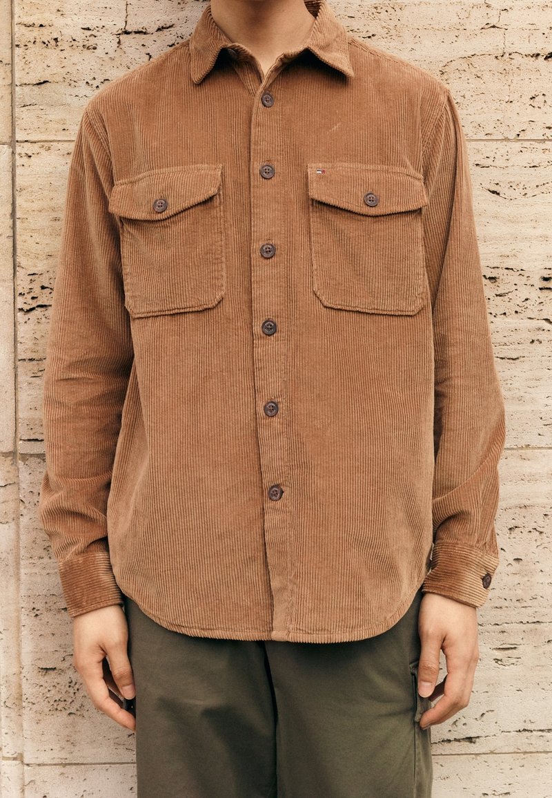 Brun kordfløjls-skjorte med to brystlommer, knaplukning foran og spids krave. Lange ærmer, ribbet tekstur og rundet kant.