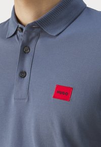 Pikétröja i mörkblått, tillverkad av textilier med struktur. Utrustad med ribbat krage, tvåknäppning och en röd rektangulär patch med "HUGO"-logotyp.