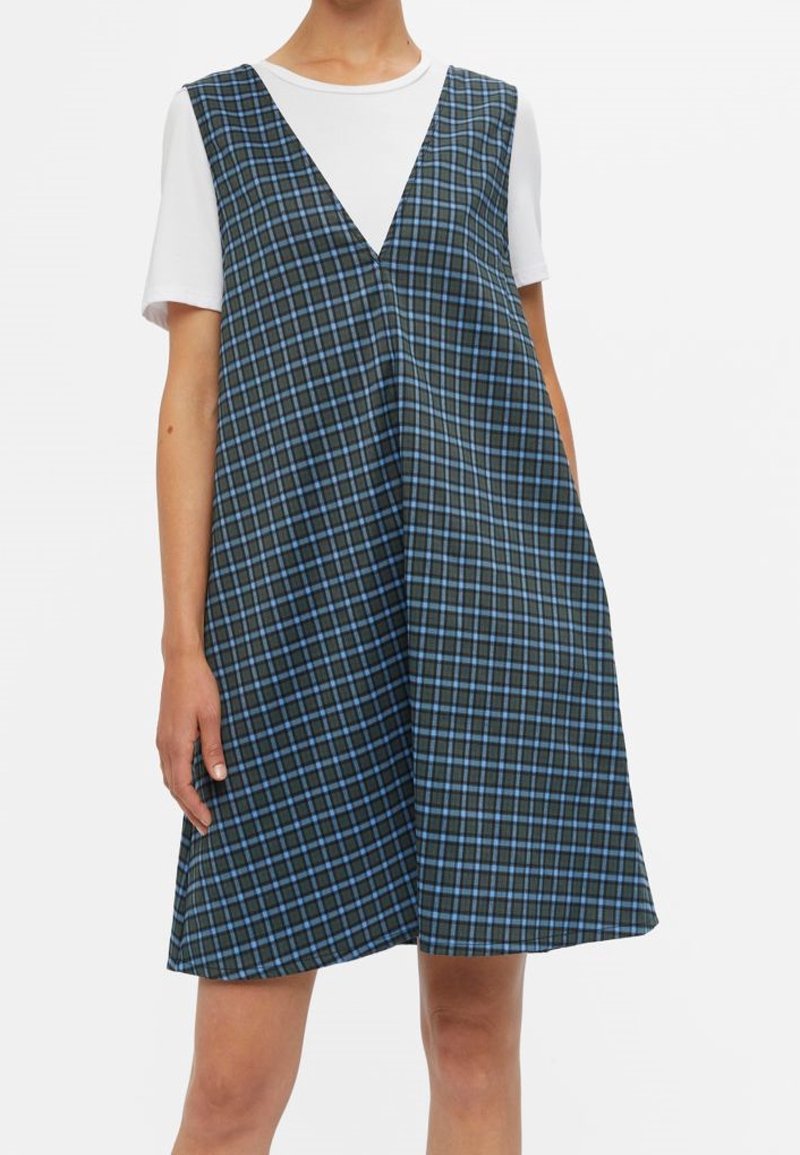 Femme portant une robe sans manches à col en V avec un motif à carreaux bleu et noir par-dessus un top blanc à manches courtes sur fond blanc.