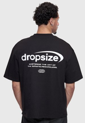 Bărbat cu păr creț, purtând un tricou negru cu text alb „dropsize” și sloganul „Mastering the art of the infinite, regardless” pe spate.