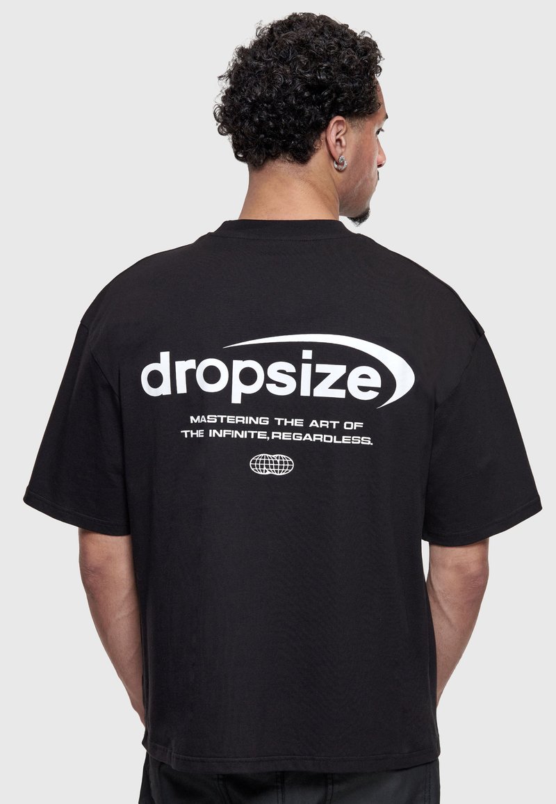 Homme aux cheveux bouclés portant un t-shirt noir avec un texte blanc « dropsize » et le slogan « Mastering the art of the infinite, regardless » dans le dos.