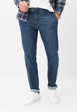 Homme portant un jean bleu retroussé, des baskets blanches, une chemise blanche et une chemise à carreaux bleue boutonnée avec une main dans la poche.