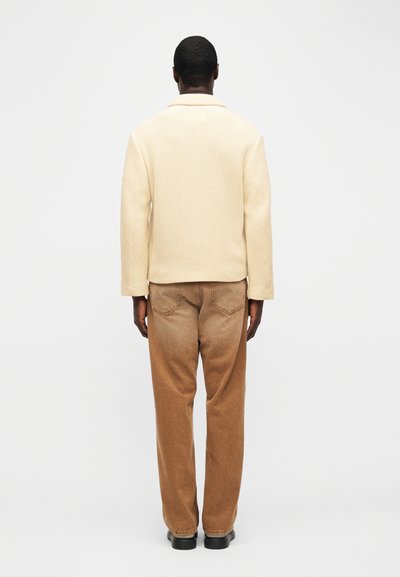 Άνδρας που στέκεται γυρισμένος με την πλάτη φοράει εκρού πλεκτό πουλόβερ, καφέ ξεβαμμένα τζιν και μαύρα παπούτσια μπροστά σε απλό λευκό φόντο.