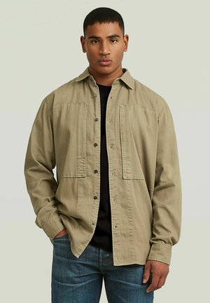 Ung mand iført beige skjorte med knapper over sort t-shirt og blå jeans, stående med den ene hånd i lommen mod en lys baggrund.