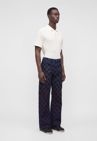 Homme debout portant un polo blanc texturé à manches courtes, un pantalon large sombre à motif et des baskets noires sur un fond uni.