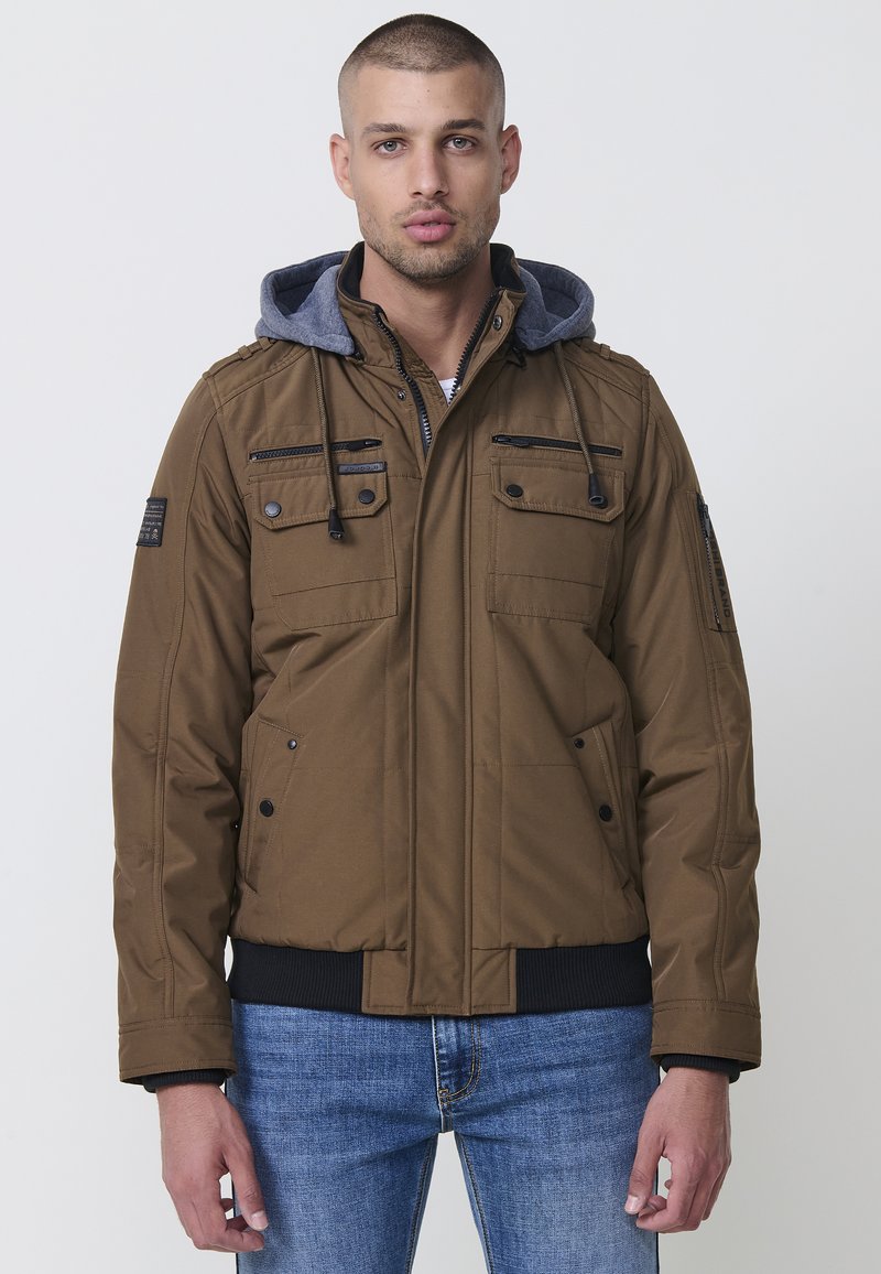 Braune isolierte Jacke mit grauer Kapuze, mit Reißverschluss geschnittenen Taschen und gerippten Bündchen. Sie hat eine glatte Textur und ein strukturiertes Design.