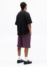 Schwarzoversized T-Shirt und lila Shorts, aus weichem Material. Die Shorts haben seitliche Taschen und einen lockeren Schnitt. Schwarze Schuhe und weiße Socken runden den Look ab.