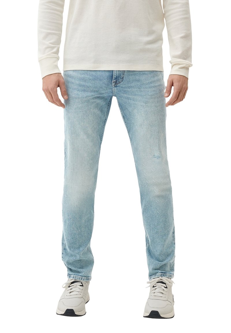 Homme portant un jean bleu clair, des baskets blanches et un chemisier crème à manches longues, debout avec les mains détendues le long du corps.