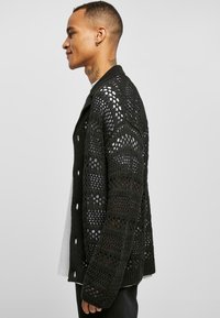 Juodas megztas kardiganas su atvirais raštais, su apykakle ir ilgomis rankovėmis. Po juo matoma šviesi apranga.