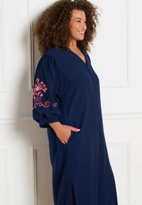 Donna con capelli ricci sorride, indossa un vestito camicia blu navy con ricamo floreale rosa sulla manica, mani nelle tasche.