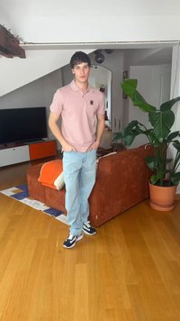 Jeune homme en polo rose et jean clair debout dans un salon avec un canapé marron, une plante en pot, une télévision et un sol en bois.