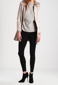 Žena v béžovém kabátu s kožešinovým límcem, šedém žebrovaném svetru, černých legínách a černých semišových kotníkových botách na vysokém podpatku, stojící.