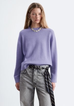 Jeune femme portant un pull lavande, un jean gris avec une ceinture foulard noire à motifs, et un collier argenté épais sur un fond blanc uni.