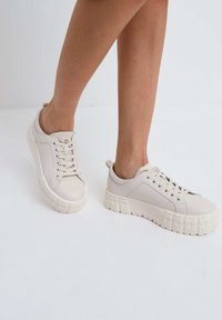 Beige sneakers met een gladde leren bovenkant, ronde neus en een gestructureerde rubberen zool met een rasterpatroon. Vetersluiting en een trekband bij de hiel.