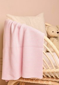 Una coperta a righe rosa e bianca adagiata su una sedia in vimini con un cuscino beige e un orsetto sullo sfondo.