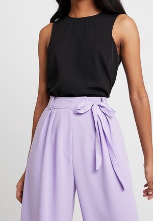 Top negro sin mangas combinado con pantalones lila de talle alto que presentan una cinturilla con lazo y un tejido texturizado, mostrando líneas limpias y bolsillos.