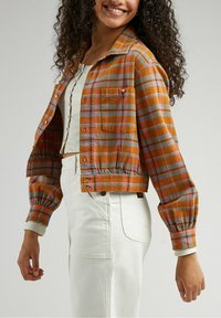 Cropped jacket in orange, blue, and grey plaid fabric; features chest pockets, button front, and elasticized cuffs; paired with white pants.